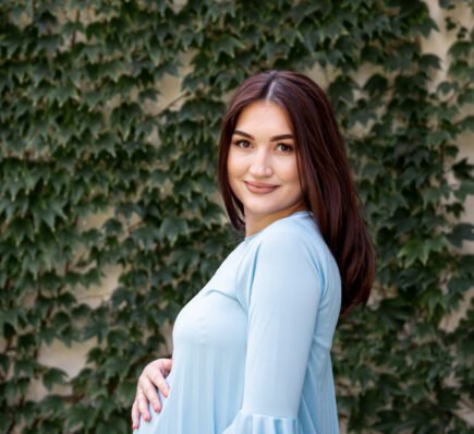 Pregnant woman in a light blue dress smiling while holding her baby bump, standing against a leafy green background.