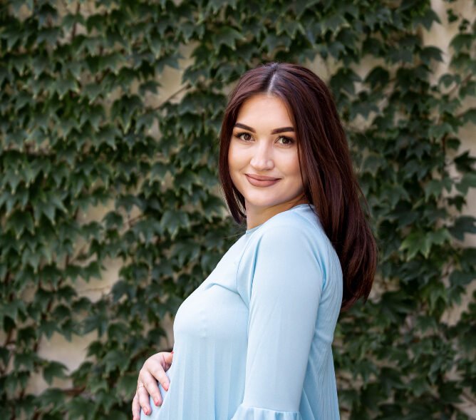 Pregnant woman in a light blue dress smiling while holding her baby bump, standing against a leafy green background.