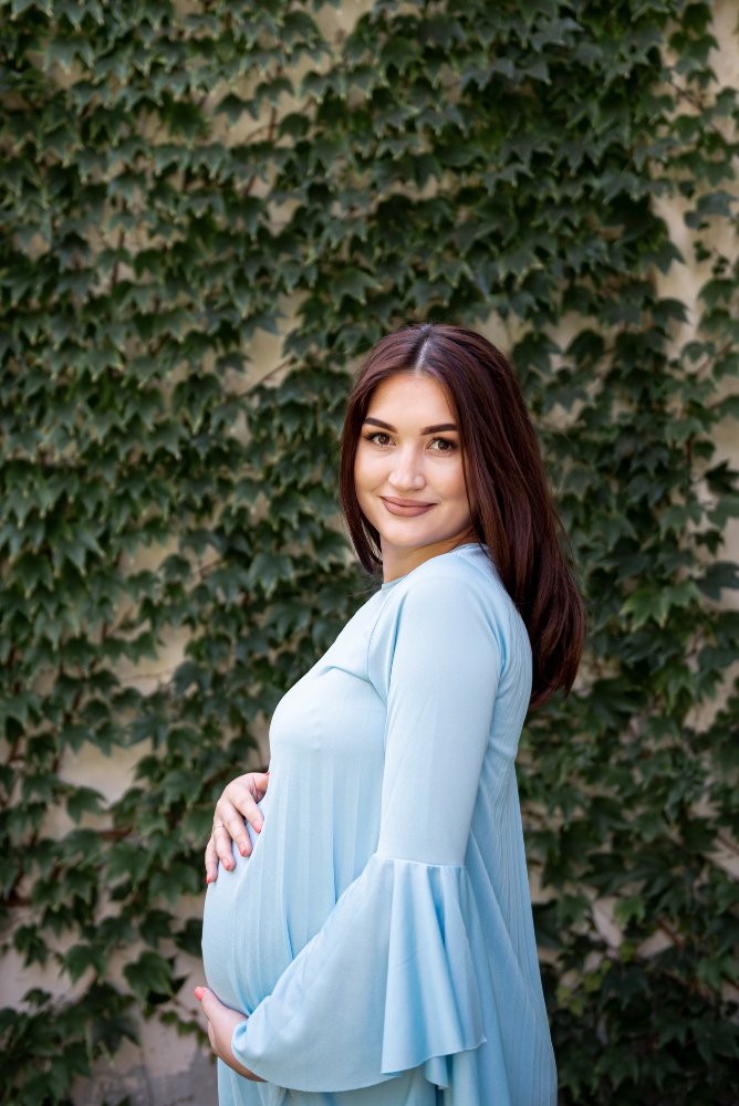 Pregnant woman in a light blue dress smiling while holding her baby bump, standing against a leafy green background.