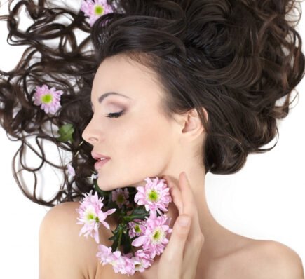 A woman with long, wavy dark hair lies gracefully with her eyes closed, holding pink flowers near her face, while blossoms are also woven into her hair.