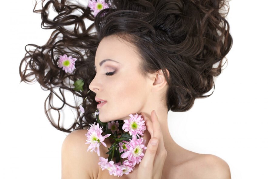A woman with long, wavy dark hair lies gracefully with her eyes closed, holding pink flowers near her face, while blossoms are also woven into her hair.