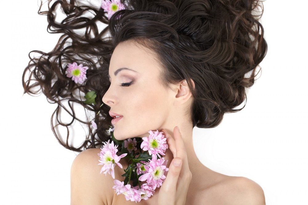 A woman with long, wavy dark hair lies gracefully with her eyes closed, holding pink flowers near her face, while blossoms are also woven into her hair.