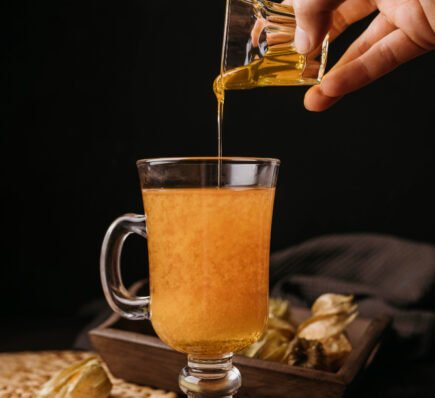 A hand pouring golden honey into a glass mug of warm drink surrounded by cinnamon sticks, star anise, and physalis fruits.