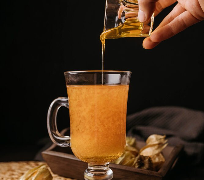A hand pouring golden honey into a glass mug of warm drink surrounded by cinnamon sticks, star anise, and physalis fruits.