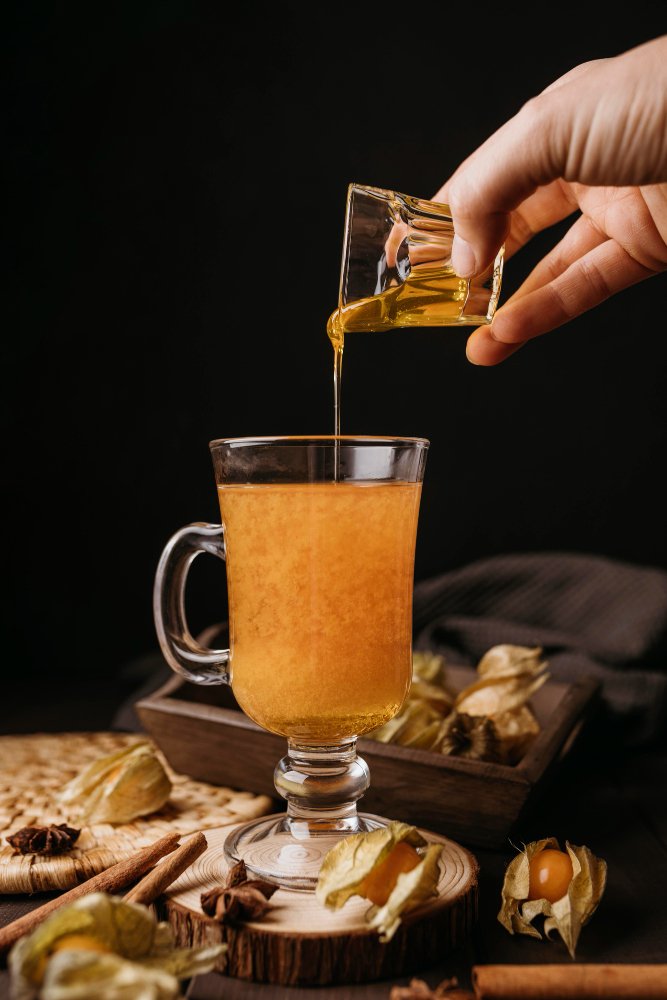 A hand pouring golden honey into a glass mug of warm drink surrounded by cinnamon sticks, star anise, and physalis fruits.