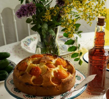 Homemade apricot cake on a decorative stand, surrounded by wildflowers, cucumbers, and a bottle of amber liquid on a lace-covered table.