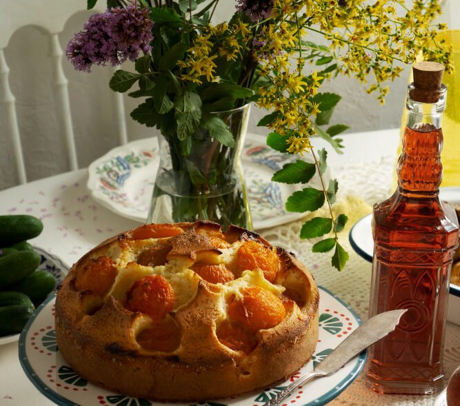 Homemade apricot cake on a decorative stand, surrounded by wildflowers, cucumbers, and a bottle of amber liquid on a lace-covered table.