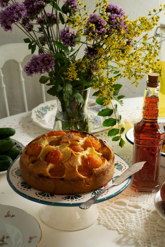 Homemade apricot cake on a decorative stand, surrounded by wildflowers, cucumbers, and a bottle of amber liquid on a lace-covered table.