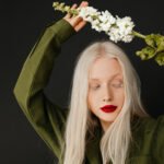 A person with platinum blonde hair and red lipstick poses with closed eyes, holding a stem of white flowers above their head against a black background.