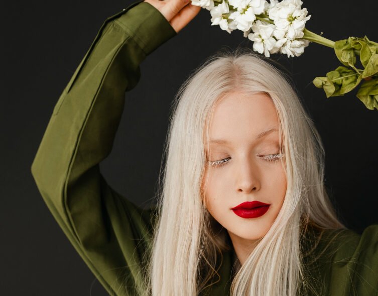 A person with platinum blonde hair and red lipstick poses with closed eyes, holding a stem of white flowers above their head against a black background.