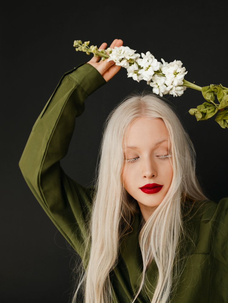 A person with platinum blonde hair and red lipstick poses with closed eyes, holding a stem of white flowers above their head against a black background.