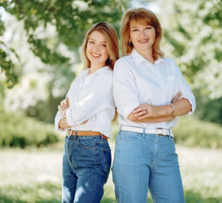 Two women standing side by side outdoors, wearing matching white shirts and blue jeans, smiling with arms crossed in a lush green park.