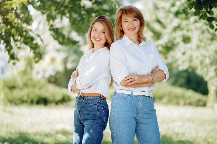 Two women standing side by side outdoors, wearing matching white shirts and blue jeans, smiling with arms crossed in a lush green park.