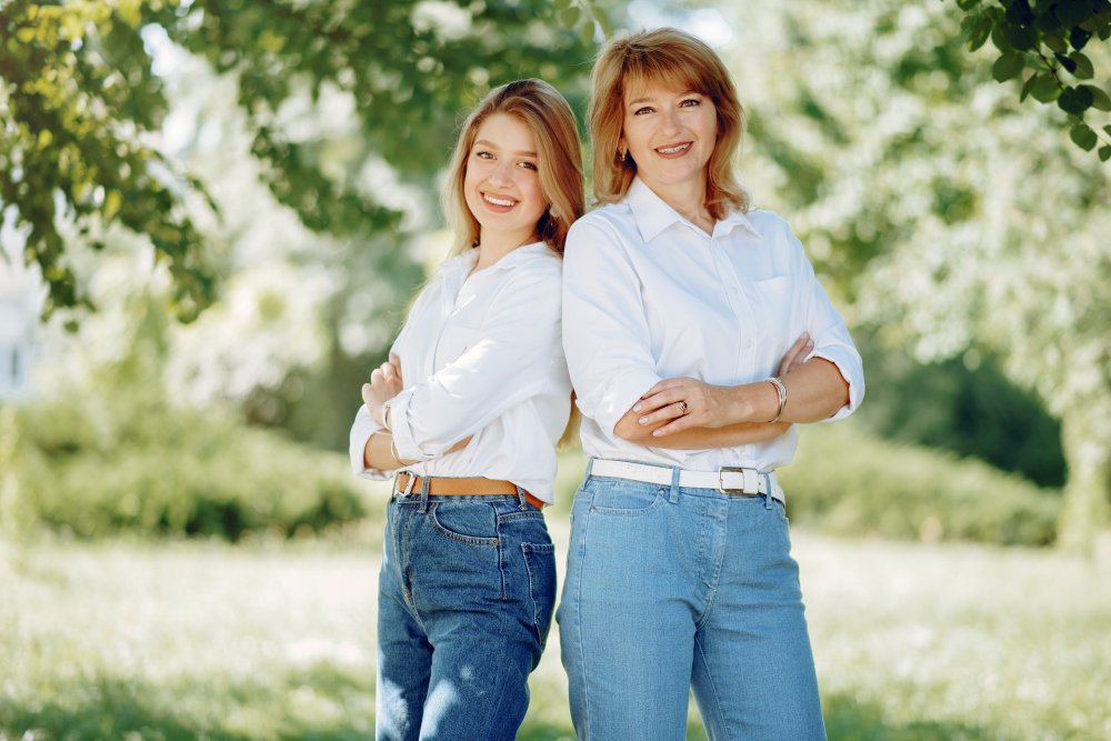 Two women standing side by side outdoors, wearing matching white shirts and blue jeans, smiling with arms crossed in a lush green park.
