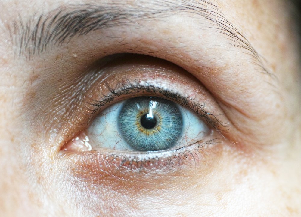 Close-up of a human eye with a vivid blue iris featuring green and yellow hues near the pupil, surrounded by visible red veins in the sclera and detailed skin texture.