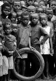 A group of African children standing closely together, some barefoot, with a worn-out tire in front of them, looking directly at the camera.