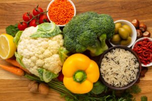 A colorful assortment of fresh, healthy foods including cauliflower, broccoli, yellow bell pepper, cherry tomatoes, lemon, carrots, walnuts, hazelnuts, green olives, red lentils, goji berries, and mixed rice, arranged on a wooden surface.