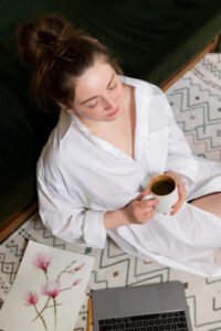 A person in a white shirt sits on a patterned rug beside a green couch, holding a cup and facing an open laptop, with a floral art print nearby
