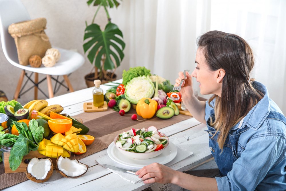 Woman enjoying a fresh vegetable salad at a white table surrounded by colorful fruits and vegetables.