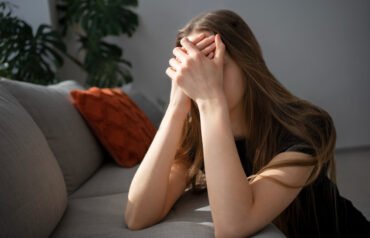 A person with long brown hair sits on a light gray couch, head in hands, face obscured. They wear a black shirt. An orange pillow and green plant are visible in a softly lit, modern indoor setting.