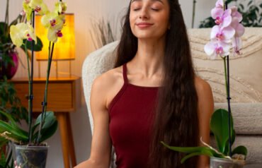 Person sitting cross-legged on a carpeted floor, peacefully holding yellow and pink orchid plants, surrounded by cozy indoor decor.