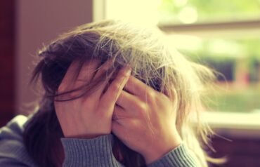 A person with long hair sits indoors near a window, wearing a ribbed sweater, with both hands covering their face and fingers tangled in their hair, conveying distress or overwhelm.