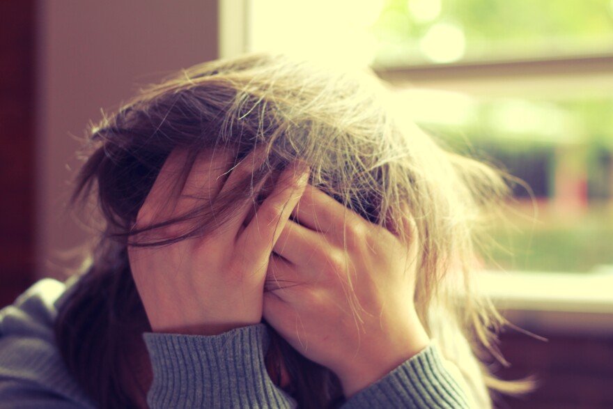 A person with long hair sits indoors near a window, wearing a ribbed sweater, with both hands covering their face and fingers tangled in their hair, conveying distress or overwhelm.
