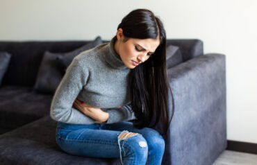 A person sitting on a dark gray couch, wearing a gray turtleneck sweater and ripped blue jeans, clutching their abdomen with both hands, suggesting discomfort or stomach pain.