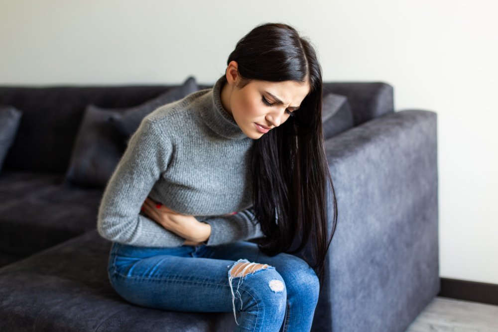 A person sitting on a dark gray couch, wearing a gray turtleneck sweater and ripped blue jeans, clutching their abdomen with both hands, suggesting discomfort or stomach pain.