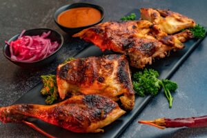 A close-up shot of grilled, charred chicken pieces served on a long black rectangular platter. The chicken is garnished with fresh parsley and accompanied by two small black bowls—one filled with red chili sauce and the other with pickled red onions—set against a dark textured background.